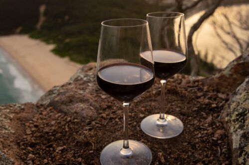 Zwei Weingläser mit australischem Rotwein auf einem Felsen mit Blick auf Strand und Meer bei Sonnenuntergang, ideal für romantische Abende.