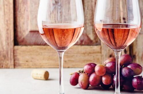 Zwei Gläser trockener Roséwein vor Holzhintergrund, begleitet von Weintrauben und einem Korken. Genießen Sie die sommerliche Frische eines edlen Tropfens!