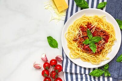 Ein Teller Spaghetti Bolognese, umgeben von Basilikumblättern, Kirschtomaten und Knoblauch, idealer Weinbegleiter zu Pasta.