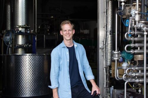 Ein Mann in blauer Hemdjacke steht vor technischen Weinausrüstungen und lächelt. Leidenschaft für alkoholfreien Wein!