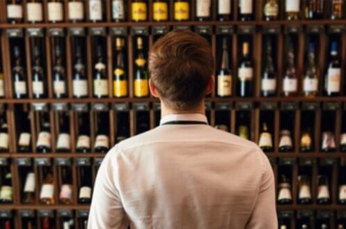 Mann in weißem Hemd steht vor einem Weinschrank mit verschiedenen Weinflaschen in einem Weinladen - entdecken Sie unsere Vielfalt an hochwertigen Weinen!