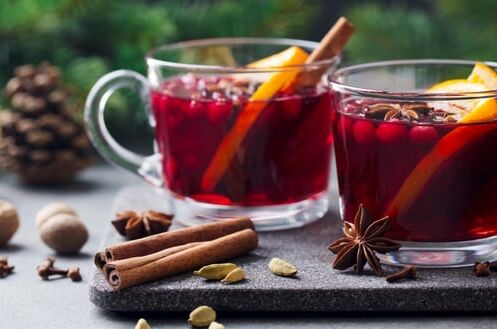 Zwei Gläser Glühwein mit Zimtstangen und Orangenscheiben, dekoriert mit Gewürzen und Tannenzapfen, auf schwarzem Untersetzer.