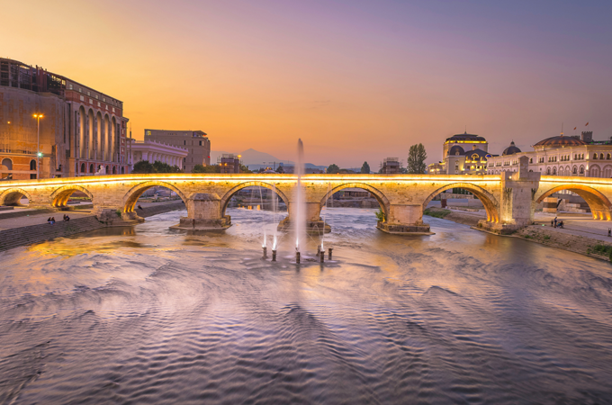 Die historische Steinbrücke in Skopje, Nordmazedonien, beleuchtet bei Sonnenuntergang über dem Fluss Vardar. Der sanft fließende Fluss reflektiert die Lichter der Stadt, während der Himmel in warmen Orange- und Lilatönen leuchtet.