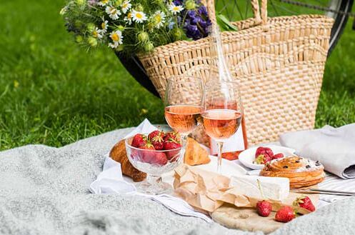 Picknickdecke mit Früchten, Croissants und Käse, zwei Gläser Roséwein, Fahrrad im Hintergrund, Frühling.