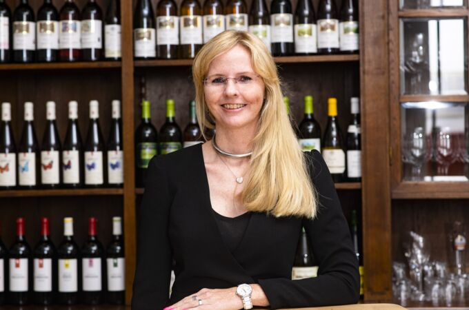 Blonde Frau im schwarzen Kleid vor einem Weinregal. Sie trägt eine Uhr und lächelt.