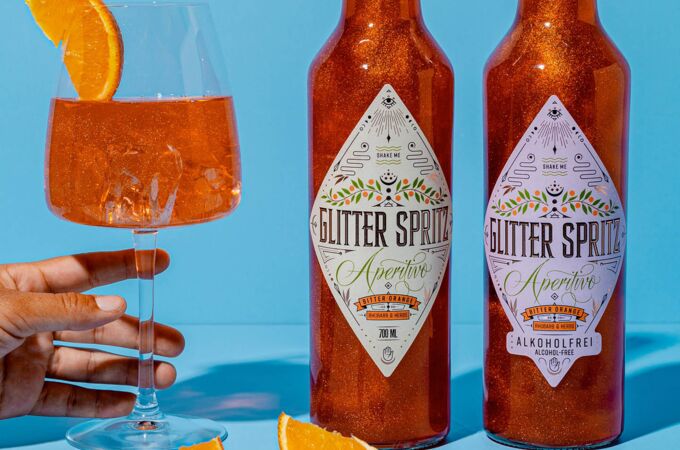 Craft Circus Glitter Spritz-Flaschen und ein Glas mit Orangenscheibe vor blauem Hintergrund. Erfrischender Aperitif mit Glitzereffekt und sattem Orangengeschmack.