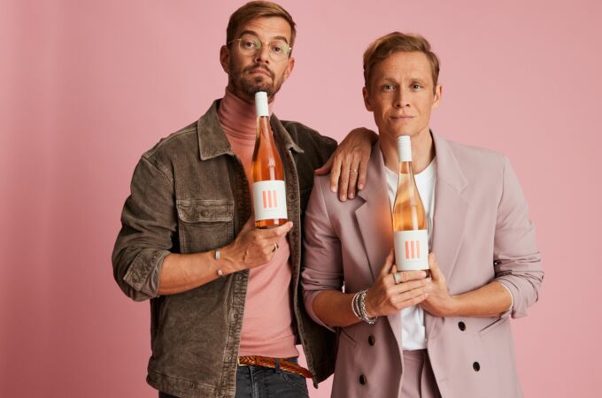 Zwei Männer halten jeweils eine Flasche III FREUNDE Wein in den Händen. Beide tragen stylische Outfits und posieren vor einem rosa Hintergrund. Hochwertige deutsche Weine, perfekt für besondere Anlässe.