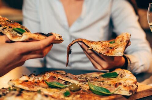 Zwei Personen halten Stücke von Pizza in den Händen, serviert mit einem Glas Weißwein auf einem Tisch, ideal für gemütliche Abende.