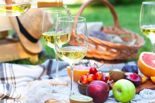 Ein Picknick im Frühling mit Obst und drei Gläsern Weißwein, die auf einer Decke eingeschenkt werden.