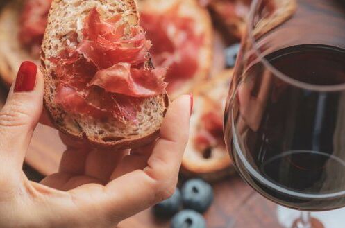 Eine Hand hält ein Stück Brot mit spanischem Schinken, daneben ein Glas trockener spanischer Rotwein.