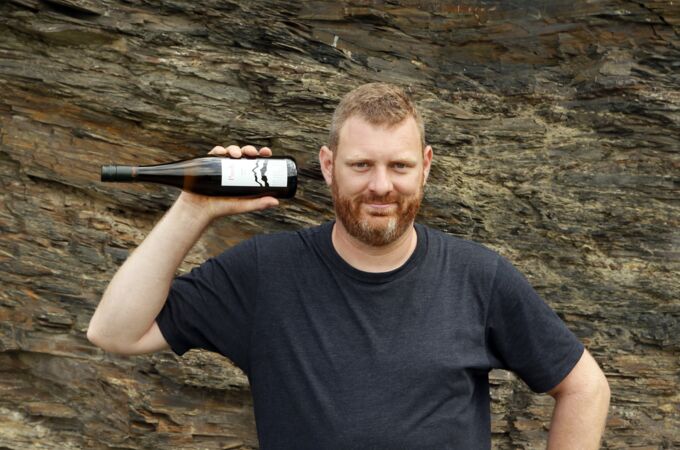 Mann in schwarzem T-Shirt hält eine Flasche Axel Pauly Wein vor einer steinigen Wand. Ideal für Weinliebhaber!