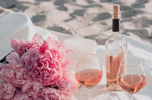 Zwei Gläser italienischer Roséwein und Blumenstrauß mit Sonnenhut auf Sand am Strand, Sommer-Ambiente.