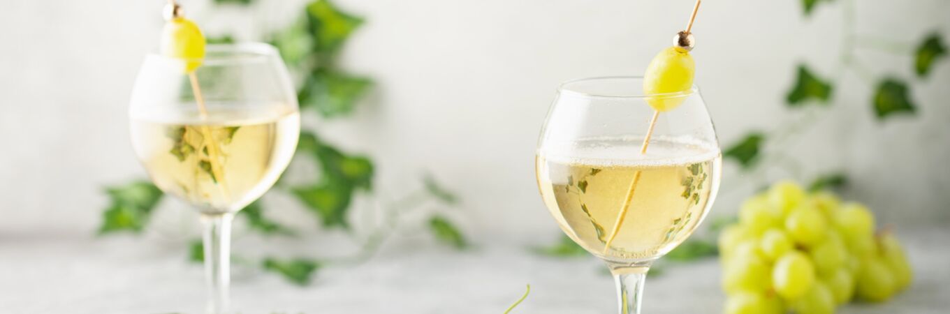 Ein Glas Weinschorle mit weißem Wein und Traube, serviert in elegantem Glas mit frischen Trauben und Weinblättern im Hintergrund.