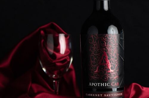 Flasche Apothic Cab Cabernet Sauvignon Rotwein vor elegantem roten Tuch und leeren Weinglas, dunkler Hintergrund. Genießen Sie luxuriösen Geschmack!