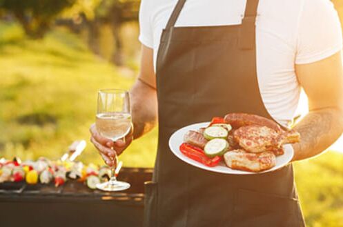 Mann im Freien mit weißem Hemd und dunkler Schürze hält ein Glas Weißwein und einen Teller mit Grillfleisch und Gemüse.