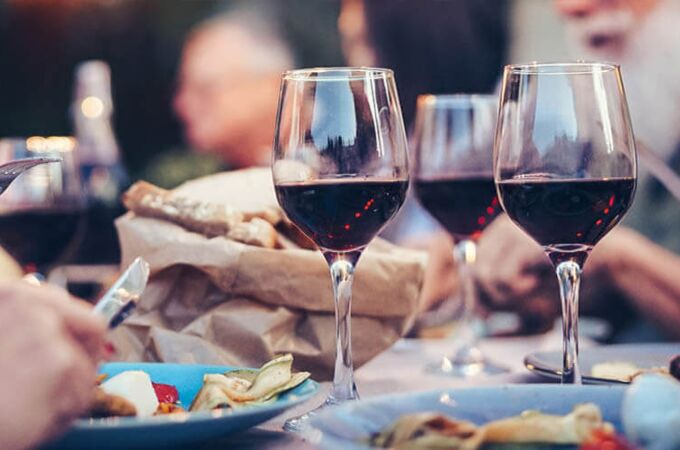 Zwei Gläser Malbec-Wein auf einem gedeckten Tisch bei einem Essen im Freien, umgeben von Personen, die speisen.