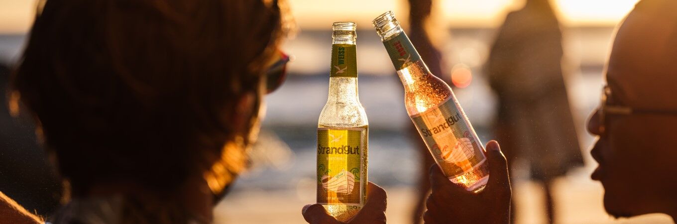 Strandgut Weinschorle Zwei Personen genießen Strandgut Weinschorle in Weiß und Rosé bei Sonnenuntergang am Strand – ready to drink, leicht und perfekt für den Moment.