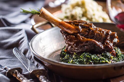 Schmackhaftes Osterlamm in Edelstahlschüssel mit grünem Gemüse, rotem Wein und grau gedecktem Tisch. Perfekte Kombination aus Wein und Essen.