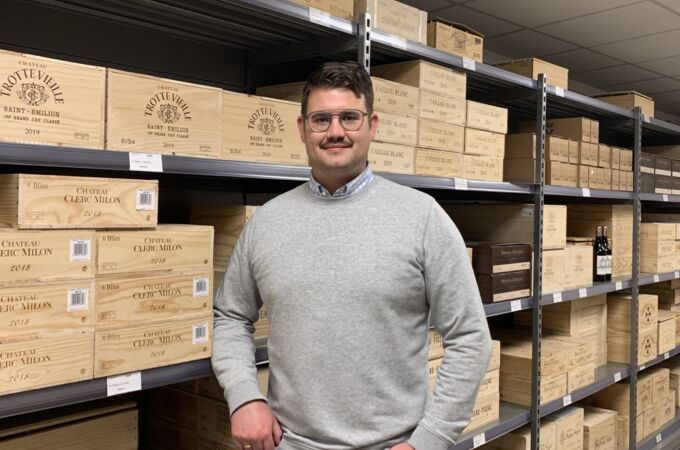 Ein Mann im grauen Pullover steht vor Regalen mit Holzkisten von Weingütern wie Chateau Clerc Milon und Chateau Trotte Vieille.