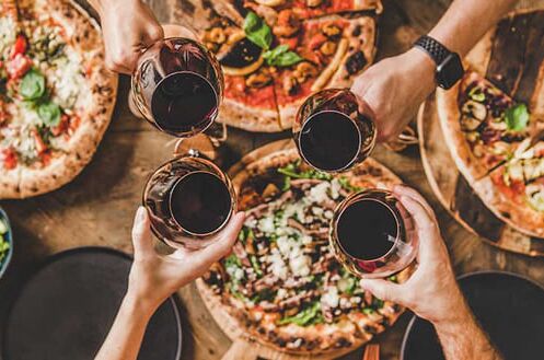 Vier Personen stoßen mit Rotweingläsern über einem Tisch voller Pizzen und einem Salat an, Rotwein und Pizza genießen.