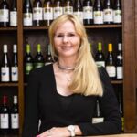 Blonde Frau im schwarzen Kleid vor einem Weinregal. Sie trägt eine Uhr und lächelt.