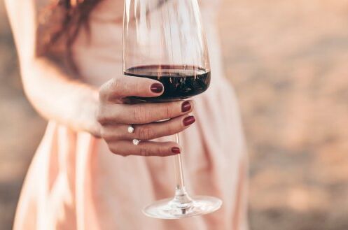 Frau in zartem Kleid hält portugiesischen Wein im Glas vor ländlicher Kulisse. Dunkelroter Wein im klaren Glas.