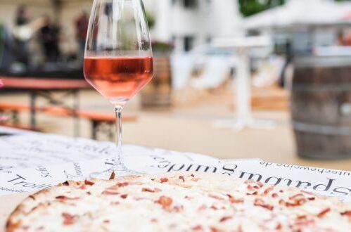 Ein Glas Roséwein und Flammkuchen auf einem Tisch im Freien, im Hintergrund eine Weinfass-Dekoration. Perfekt für gemütliche Abende im Freien!