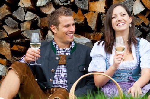 Ein lächelndes Paar in traditioneller Kleidung genießt Grünen Veltliner vor einem Stapel Holzscheite. Der perfekte Weinmoment!