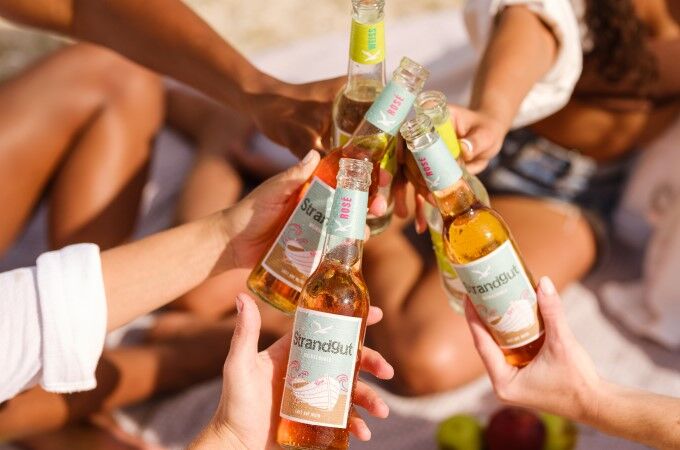 Strandgut Weinschorle unterwegs Menschen stoßen mit Strandgut Weinschorle in Weiß und Rosé an – sommerliche Szene am Strand mit ready-to-drink Schorleflaschen in geselliger Runde.