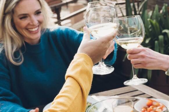 Drei lachende Frauen stoßen mit Weißwein-Gläsern auf einer Terrasse an, umgeben von Pflanzen und einem gedeckten Tisch.