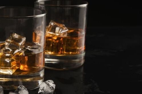 Zwei Gläser Whisky mit Eiswürfeln auf einem schwarzen Untergrund, daneben lose Eiswürfel. Perfekt als Geschenkset.