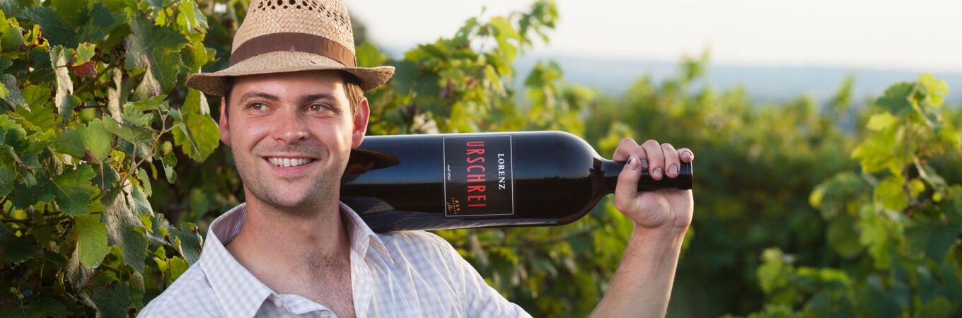 Ein Winzer vom Bioweingut Lorenz im Weinberg trägt eine Flasche Urschrei-Wein auf der Schulter, mit grünen Reben im Hintergrund - Qualität in jedem Schluck!