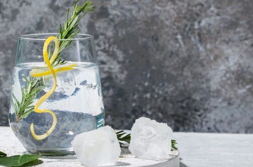 Ein Glas alkoholfreier Gin mit Rosmarinzweig und Zitronenschleife, umgeben von Eiswürfeln, auf einem grauen Hintergrund. Perfekt für erfrischende Momente!