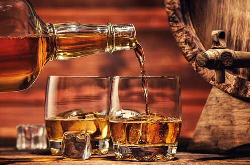 Eine Flasche amerikanischer Whiskey wird in zwei Gläser mit Eiswürfeln neben einem Holzfass eingeschenkt. Genießen Sie authentischen Geschmack.