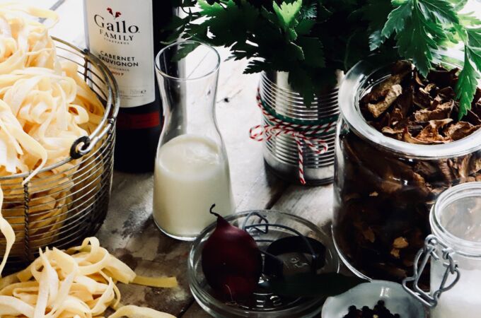 Eine Flasche Gallo Wein Cabernet Sauvignon steht auf einem Holztisch, umgeben von frischer Pasta, einem Bündel Petersilie, Sahne in einer Karaffe, getrockneten Pilzen und Gewürzen.