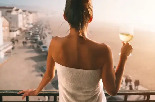 Eine Frau in einem weißen Handtuch hält ein Glas portugiesischen Weißwein und genießt den Sonnenuntergang von einem Balkon mit Meerblick.