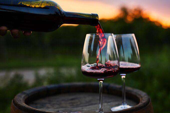 Ein Glas roter Bordeaux-Wein wird aus einer Flasche in zwei elegante Weingläser auf einem rustikalen Holzfass eingeschenkt. Im Hintergrund ist eine verschwommene, grüne Landschaft und ein stimmungsvoller Sonnenuntergang zu sehen.