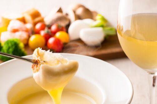 Ein Glas Weißwein und eine Gabel mit Brot, getunkt in Käsefondue, mit frischen Zutaten im Hintergrund. Ideal zu Ihrer Käsefondue-Mahlzeit!