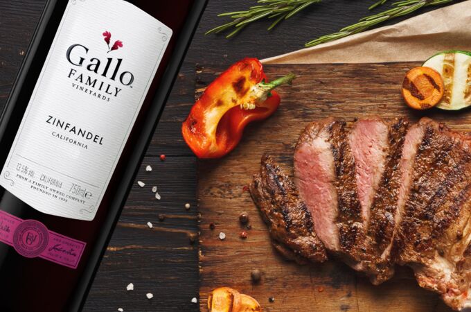 Eine Flasche Gallo Zinfandel Rotwein neben einem Holzbrett mit gegrilltem, in Scheiben geschnittenem Steak, gegrilltem Gemüse, Rosmarinzweig und Gewürzen.