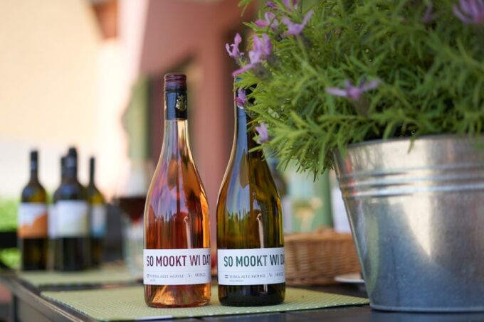 Flaschen Roséwein und Weißwein der Marke Montigny SO MOOKT WI DAT im Freien, flankiert von Blumen, perfekt für entspannte Sommerabende.