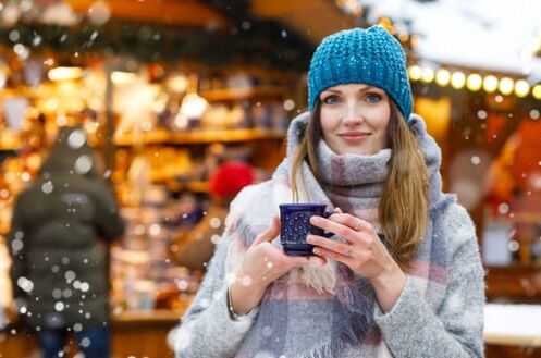 Eine Frau genießt auf einem verschneiten Weihnachtsmarkt Glühwein aus einer Tasse. Perfektes Getränk für kalte Wintertage!