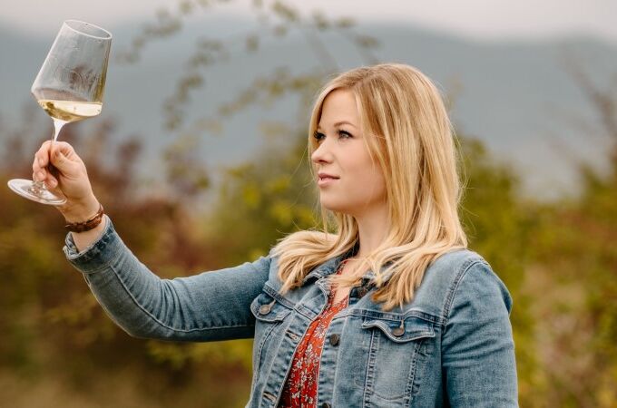 Eine Frau in Jeansjacke begutachtet ein Glas Weißwein im Freien, markiert durch klare Reflexionen. Genießer Moment in den Weinbergen.