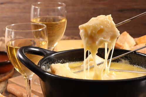 Fondue-Set mit zwei Gläsern Weißwein und Baguette auf Holzbrett. Perfekt für den Genuss an Silvester!