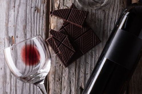 Flasche süßer Rotwein mit leerem Weinglas und dunkler Schokolade auf rustikalem Holztisch, ideal für gemütliche Abende.