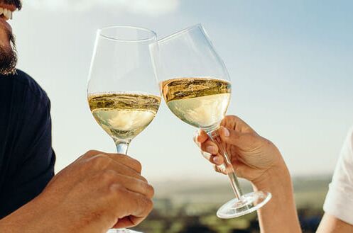 Zwei Personen stoßen mit Weingläsern voller Chardonnay bei sonnigem Wetter an, im Hintergrund eine Weinlandschaft. 