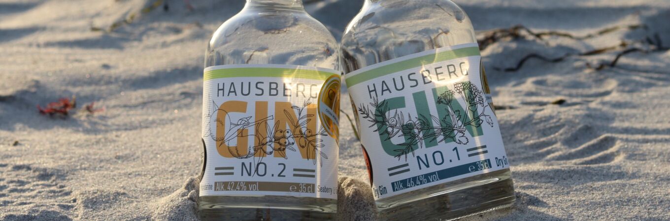 Zwei Flaschen Hausberg Gin No. 2 und No. 1 auf Sandstrand bei Sonnenuntergang. Perfekt für sommerliche Genussmomente.