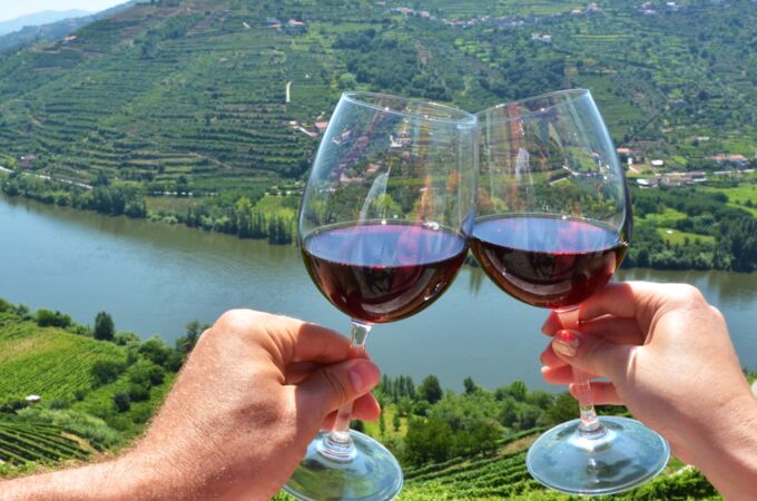 Zwei Hände halten Weingläser mit rotem Portwein im Douro-Tal, mit malerischen Weinbergen im Hintergrund. Genießen Sie exquisite Weine in idyllischer Umgebung.