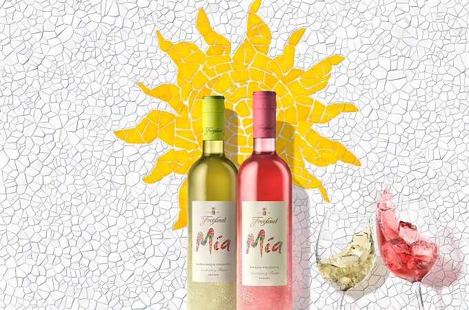 Zwei Freixenet Mia Weinflaschen, eine weiße und eine rote, vor einer bunten Mosaikwand, daneben zwei Gläser mit Wein.