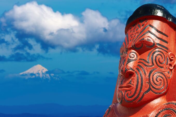 Ein kunstvoll geschnitztes, rot bemaltes Māori-Totem mit traditionellen Mustern im Vordergrund. Im Hintergrund erhebt sich ein schneebedeckter Berg unter einem klaren, blauen Himmel mit vereinzelten Wolken.