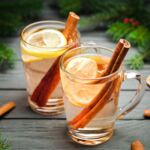 Zwei Gläser Weißwein-Glühwein mit Scheiben Zitrone und Zimtstangen, dekoriert mit Gewürzen auf Holztisch, weihnachtliche Atmosphäre.